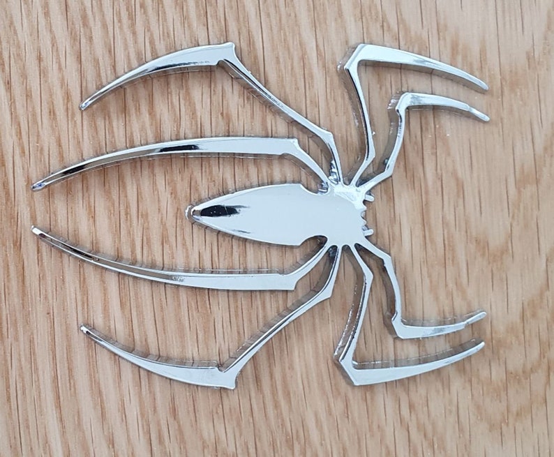 Pode incluir: Um recorte de metal de aranha prateado com oito pernas. A aranha est&aacute; sobre um fundo de madeira.