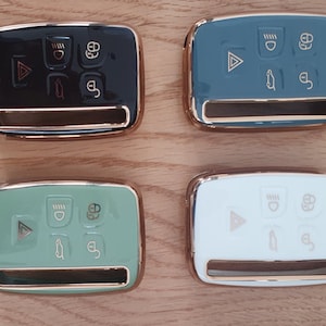 May include: Four key fobs with different coloured cases. The cases are black, teal, green, and white. Each case has a gold trim and features five buttons. The buttons are a triangle, a circle with a line through it, a car icon, a circle with a line through it, and a circle with a keyhole.