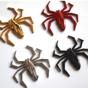 May include: Four metal spider car emblems in different colors: gold, red, silver, and black. Each spider has a lightning bolt design on its body.