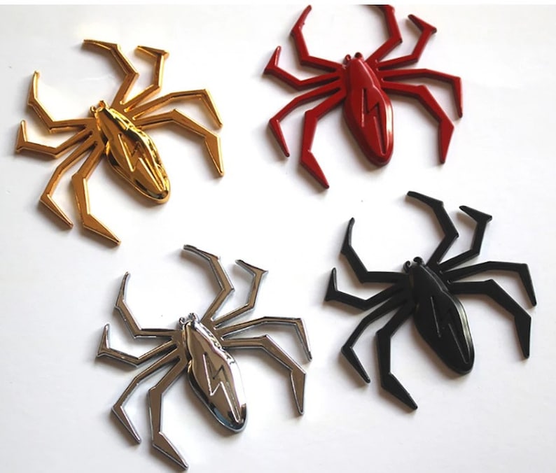May include: Four metal spider car emblems in different colors: gold, red, silver, and black. Each spider has a lightning bolt design on its body.