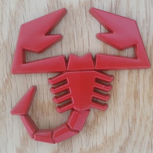 May include: A red plastic scorpion emblem with a detailed design. The scorpion has a raised tail and a segmented body.