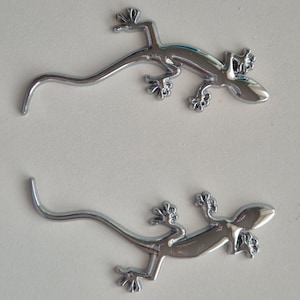 Op de afbeelding: Twee glanzende, chroomkleurige decoratieve accenten in de vorm van een hagedis. Elke hagedis heeft een lange, gebogen staart, vier gedetailleerde voeten en een glad, reflecterend lichaam. De hagedissen zijn geplaatst op een lichtgekleurd oppervlak.