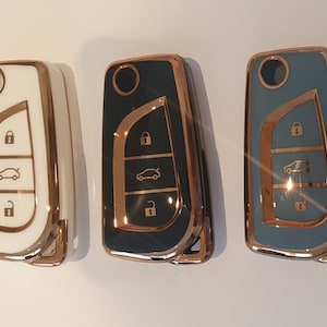 May include: Three car key fobs with gold trim. The key fobs are white, black, and gray. Each fob has three buttons and a car icon.