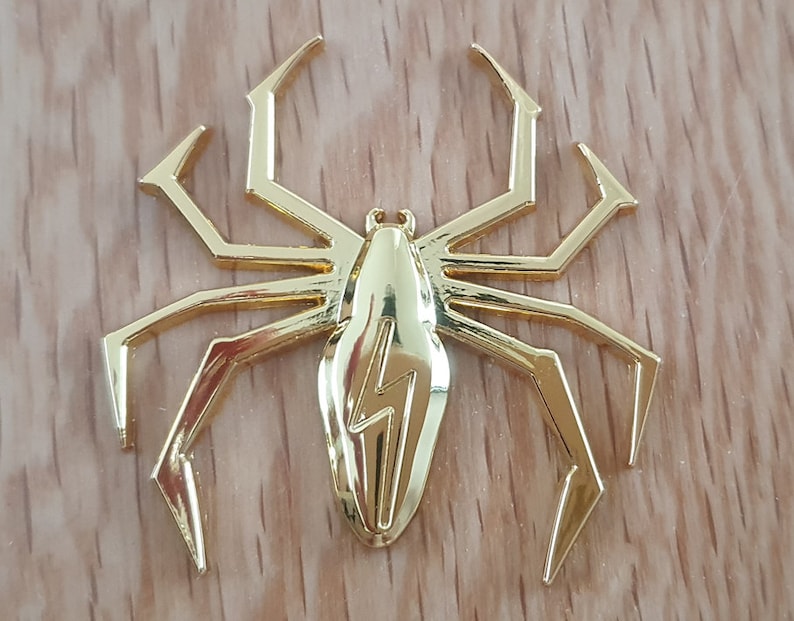May include: A gold-colored metal spider with a lightning bolt design on its body. The spider has eight legs and is a decorative piece.