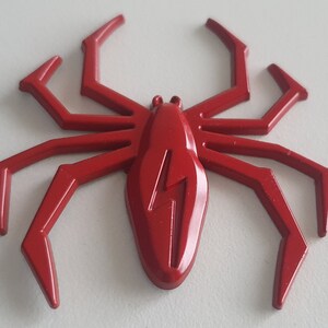 May include: A red plastic spider with a lightning bolt design on its body. The spider has eight legs and is a decorative item.