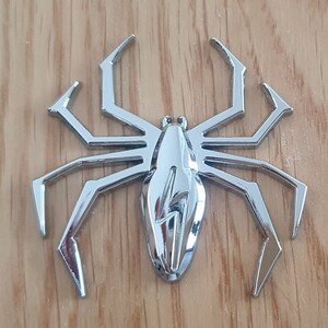 May include: A silver-colored spider-shaped car emblem with a lightning bolt design in the center.