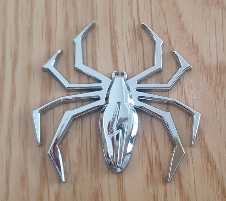 May include: A silver-colored spider-shaped car emblem with a lightning bolt design in the center.