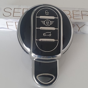 Può includere: Un portachiavi per auto nero e argento con quattro pulsanti. I pulsanti sono etichettati con icone per sbloccare, bloccare, bagagliaio e una silhouette di un'auto. Il logo Mini Cooper è visibile sul portachiavi.