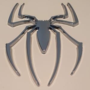 Pode incluir: Um emblema de aranha cromado com um design detalhado. A aranha est&aacute; voltada para a frente com as pernas estendidas.