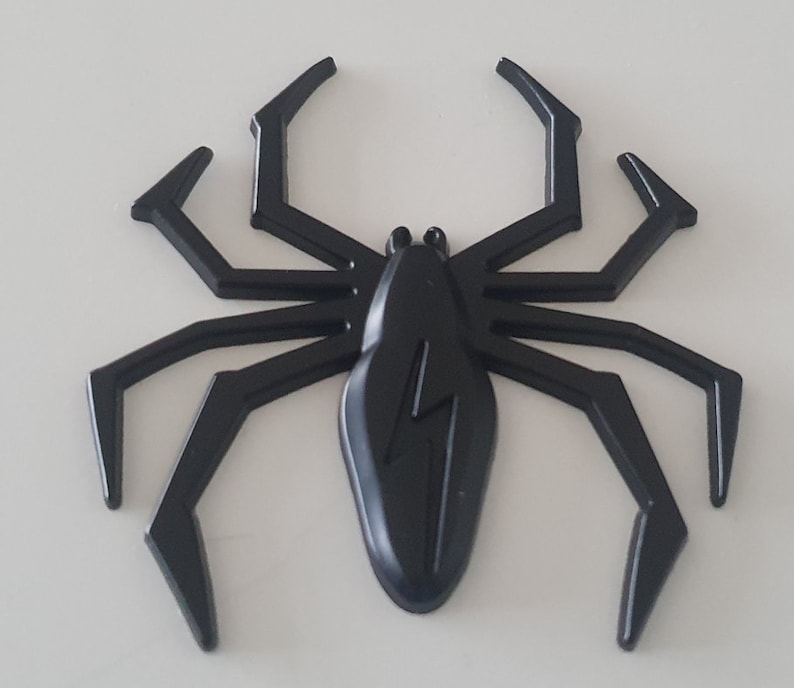 May include: Black plastic spider with a lightning bolt design on its body. The spider has eight legs and is in a raised position.