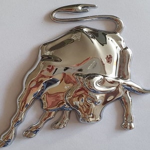 Puede incluir: Un emblema de toro cromado con un diseño en relieve tridimensional. El toro está mirando hacia la izquierda con la cabeza baja y los cuernos apuntando hacia adelante.