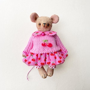 May include: A beige fabric mouse doll with dark eyes and a stitched nose. It wears a pink knitted sweater featuring a cherry appliqué, a pink collared shirt with a cherry print, and a matching pink cherry-patterned skirt over polka-dot pants.
