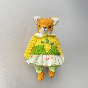 May include: A plush fox toy with orange fur and white inner ears. It wears a yellow and green knitted jumper featuring a yellow embroidered flower, a white floral print skirt with pink and orange blossoms, and light green polka dot trousers.