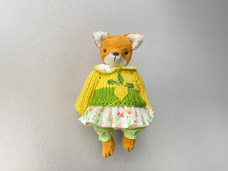 May include: A plush fox toy with orange fur and white inner ears. It wears a yellow and green knitted jumper featuring a yellow embroidered flower, a white floral print skirt with pink and orange blossoms, and light green polka dot trousers.