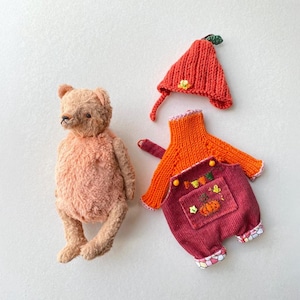 May include: A light brown plush teddy bear next to a set of doll clothing consisting of a bright orange knitted hat with a green leaf, an orange long-sleeved knitted sweater, and dark red corduroy overalls with a pumpkin embroidery.