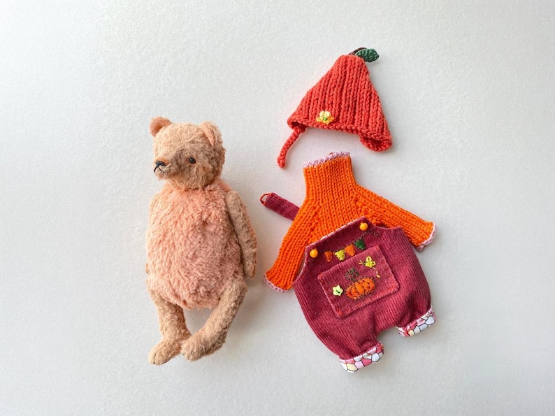 May include: A light brown plush teddy bear next to a set of doll clothing consisting of a bright orange knitted hat with a green leaf, an orange long-sleeved knitted sweater, and dark red corduroy overalls with a pumpkin embroidery.