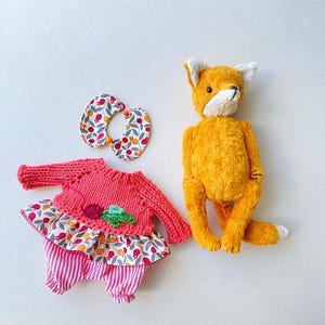 May include: A plush orange fox toy positioned next to a set of doll clothing consisting of a pink knitted sweater with a floral fabric skirt, pink and white striped trousers, and a matching floral fabric collar with a small orange bead closure.