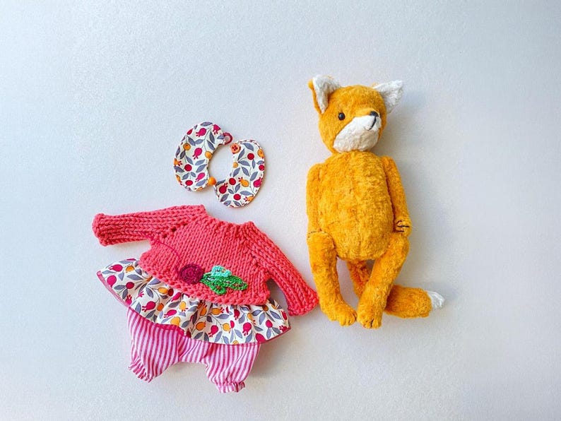 May include: A plush orange fox toy positioned next to a set of doll clothing consisting of a pink knitted sweater with a floral fabric skirt, pink and white striped trousers, and a matching floral fabric collar with a small orange bead closure.