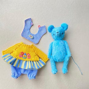 May include: A bright blue plush bear toy next to a yellow knitted long-sleeve top with three embroidered eggs, blue and yellow striped skirt trim, and blue and white gingham trousers, accompanied by a matching blue and white gingham bib.