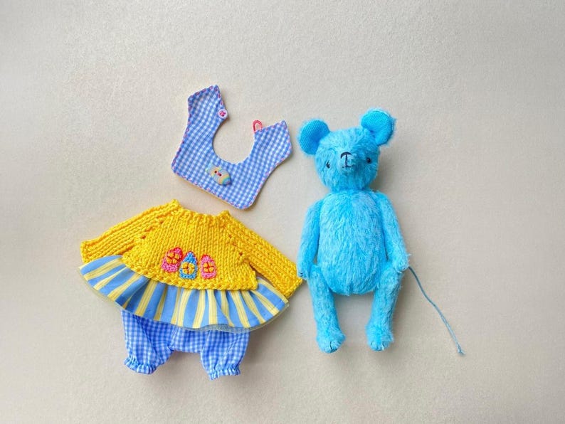 May include: A bright blue plush bear toy next to a yellow knitted long-sleeve top with three embroidered eggs, blue and yellow striped skirt trim, and blue and white gingham trousers, accompanied by a matching blue and white gingham bib.