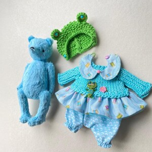 May include: A bright blue plush bear, a green crocheted frog hat with blue button eyes, and a light blue outfit consisting of a knit top with a Peter Pan collar, a small frog applique, and patterned fabric trousers with a ruffled skirt overlay.