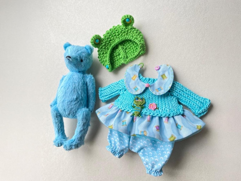 May include: A bright blue plush bear, a green crocheted frog hat with blue button eyes, and a light blue outfit consisting of a knit top with a Peter Pan collar, a small frog applique, and patterned fabric trousers with a ruffled skirt overlay.