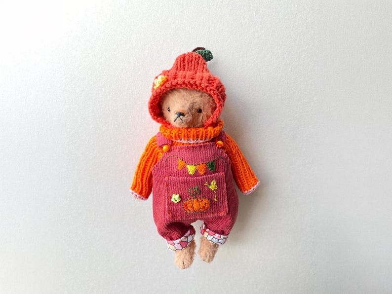 May include: A small teddy bear wearing a bright orange knitted jumper and a matching pumpkin-shaped hat. The bear is dressed in dark pink corduroy dungarees featuring embroidered pumpkin and leaf motifs on the front pocket and cuffs.