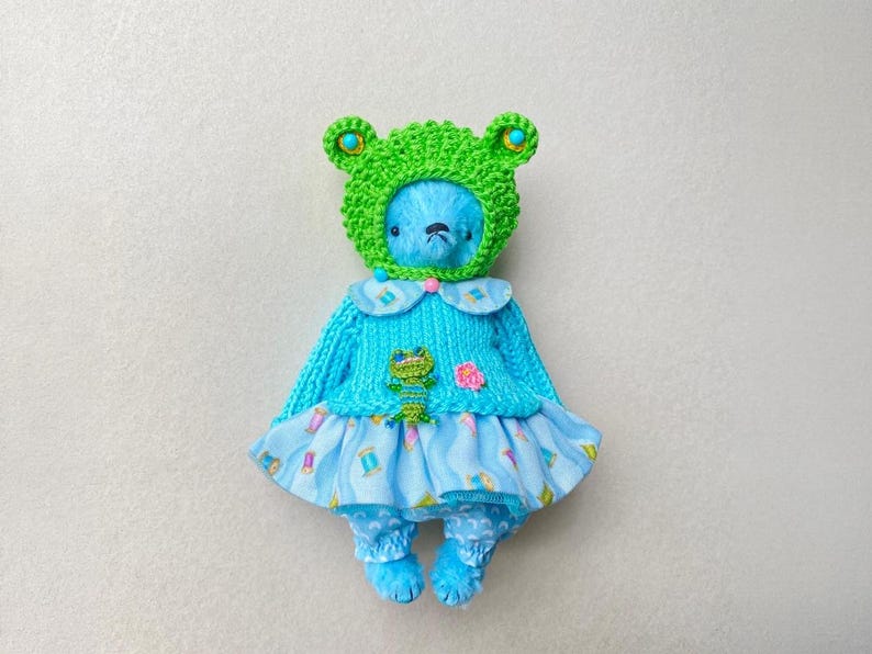 May include: A blue plush bear wearing a bright green crocheted frog hat with small turquoise accents. The bear is dressed in a light blue knitted jumper featuring a small green frog applique and a pink flower, paired with a light blue patterned skirt.