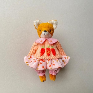 May include: A plush fox toy with orange fur and white inner ears, wearing a peach-coloured knitted sweater featuring three embroidered orange carrots, a pink and white striped collar, and a pink skirt with a small orange pattern over matching trousers.