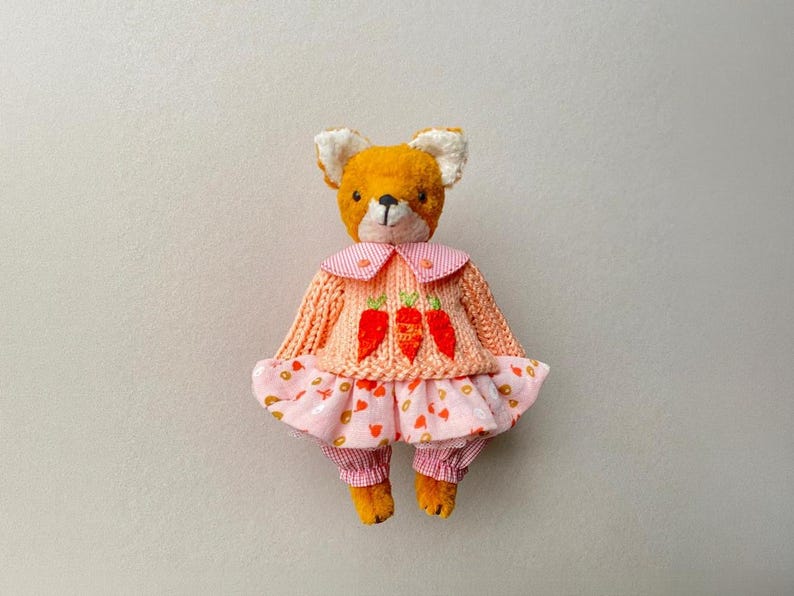 May include: A plush fox toy with orange fur and white inner ears, wearing a peach-coloured knitted sweater featuring three embroidered orange carrots, a pink and white striped collar, and a pink skirt with a small orange pattern over matching trousers.