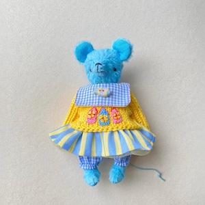 May include: A blue plush mouse toy wearing a yellow knitted jumper with three small embroidered house motifs, a blue and white gingham collar, and a blue and yellow striped skirt over blue and white checked trousers with a thin blue tail.
