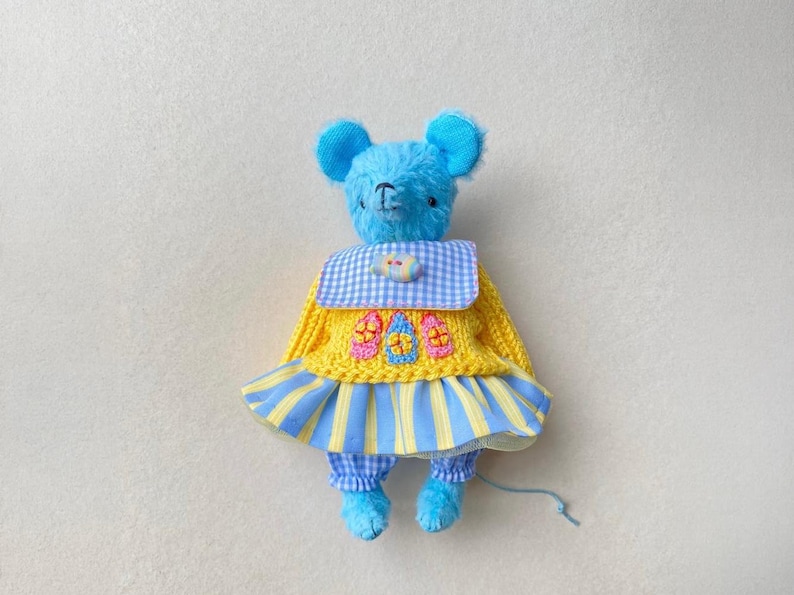 May include: A blue plush mouse toy wearing a yellow knitted jumper with three small embroidered house motifs, a blue and white gingham collar, and a blue and yellow striped skirt over blue and white checked trousers with a thin blue tail.
