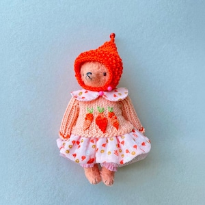May include: A small plush bear toy wearing a bright orange knitted hood, a peach-coloured sweater featuring three embroidered orange carrots, and a light pink skirt patterned with small colourful shapes, finished with a ruffled hem and pink trim.