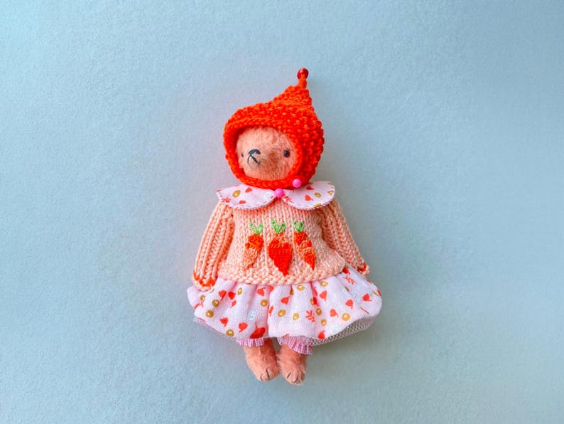 May include: A small plush bear toy wearing a bright orange knitted hood, a peach-coloured sweater featuring three embroidered orange carrots, and a light pink skirt patterned with small colourful shapes, finished with a ruffled hem and pink trim.