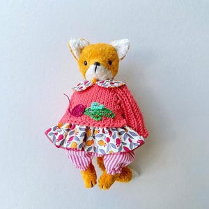 May include: A plush fox toy with orange fur, wearing a pink knitted sweater featuring a small embroidered radish, a floral patterned skirt with red and yellow motifs, and pink and white striped trousers with a matching floral collar.