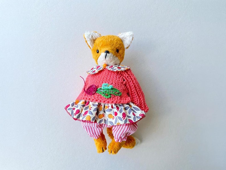 May include: A plush fox toy with orange fur, wearing a pink knitted sweater featuring a small embroidered radish, a floral patterned skirt with red and yellow motifs, and pink and white striped trousers with a matching floral collar.
