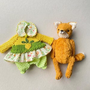 May include: A plush orange fox toy with white ear tips and a black nose positioned next to a set of doll clothing consisting of a yellow knitted jumper with a green accent, a floral print collar, and light green polka dot trousers.