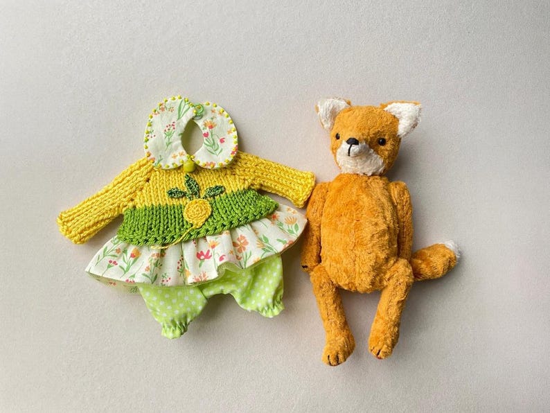 May include: A plush orange fox toy with white ear tips and a black nose positioned next to a set of doll clothing consisting of a yellow knitted jumper with a green accent, a floral print collar, and light green polka dot trousers.