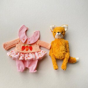 May include: A plush orange fox toy with white-tipped ears and tail, positioned next to a doll outfit consisting of a peach-coloured knit sweater with three embroidered orange carrots, a pink gingham collar, and a matching pink gingham skirt and trousers.
