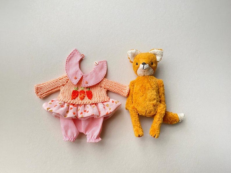 May include: A plush orange fox toy with white-tipped ears and tail, positioned next to a doll outfit consisting of a peach-coloured knit sweater with three embroidered orange carrots, a pink gingham collar, and a matching pink gingham skirt and trousers.
