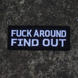 May include: A rectangular black embroidered patch featuring the white text FUCK AROUND FIND OUT in a bold, sans-serif font. The patch has a stitched border and a textured fabric surface, designed for attachment to clothing or other textile items.