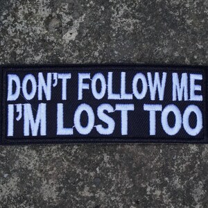 May include: A rectangular black embroidered patch featuring white stitched text that reads DON'T FOLLOW ME I'M LOST TOO in a bold, sans-serif font. The patch has a clean, finished border and is designed for application to fabric surfaces.