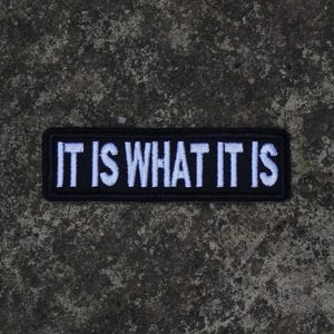 May include: A rectangular embroidered patch with a black background and a thick black border. The text IT IS WHAT IT IS is stitched in bold white capital letters across the centre of the patch, featuring a clean and legible sans-serif font style.