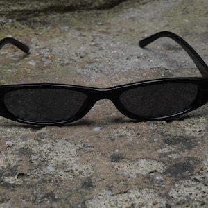 May include: A pair of black sunglasses with thin, rectangular frames and dark, tinted lenses. The glasses feature a smooth, glossy finish on the plastic frame and curved temples that extend outward from the sides of the front frame.