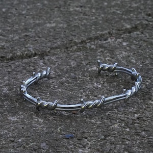 May include: A silver-toned metal cuff bracelet designed to resemble a strand of barbed wire. The bracelet features a continuous, polished metallic finish with evenly spaced, twisted spikes along the entire length of the circular, open-ended band.