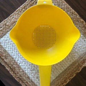 May include: A yellow plastic colander with a handle. The colander has a round bowl with a mesh bottom and a long handle.