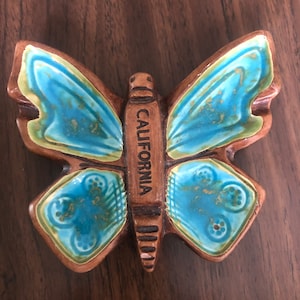 May include: A brown ceramic butterfly ashtray with blue and green glaze. The butterfly has the word "CALIFORNIA" written on its body.