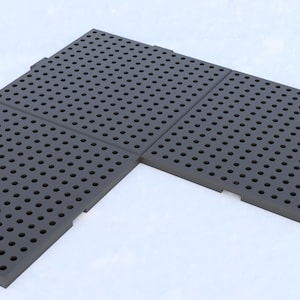 Può includere: Piastrelle per pavimenti ad incastro grigio scuro con una griglia di piccoli fori circolari. Le piastrelle sono disposte a forma di L, suggerendo un design modulare per varie applicazioni. La superficie sembra essere realizzata in un materiale resistente.