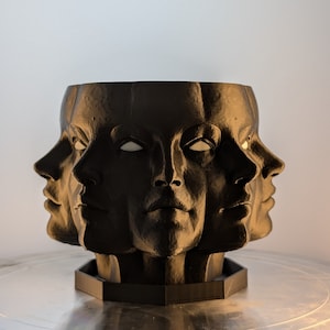 May include: A black planter shaped like a face with multiple faces surrounding it. The planter is made of a smooth, matte material and has a hexagonal base.