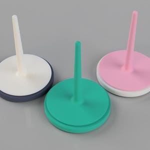 May include: Three colorful, round, ceramic pin holders. One is teal, one is pink, and one is white with a navy blue base. Each has a central, vertical, cylindrical pin. The pin holders are on a gray surface.